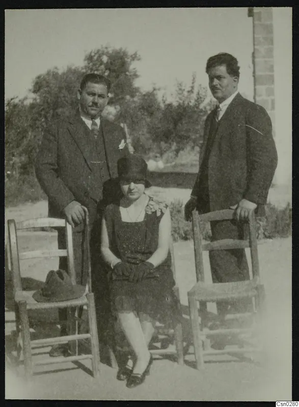 Two men in suits and a seated woman posing with wooden chairs in front of trees