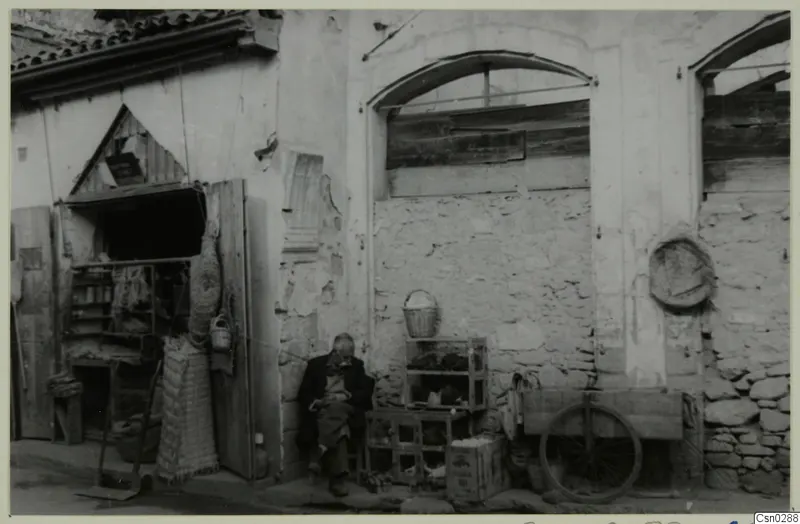 Seated man beside wire cages and a wooden cart outside a plastered building in Nicosia
