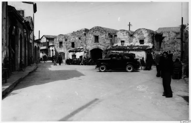 Street in Nicosia with a parked dark car, seated people, and an old stone building with arched gables behind.