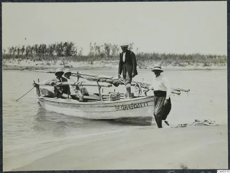 Four people with a wooden boat in shallow water at the edge of a sandy shore, reeds behind them.