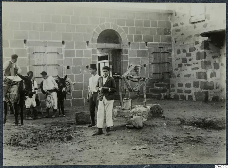 Two young men stand before a stone building with arched doorway, with two mules and three men to the left