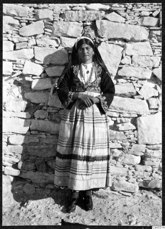 Woman in traditional dress with a dark head shawl, standing against a dry-stone wall.