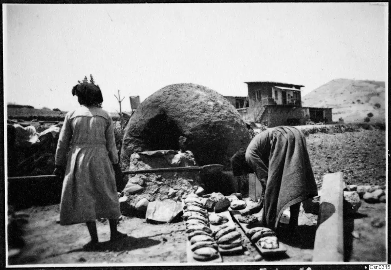 Two women at a domed clay oven, one bending to set out flatbreads on wooden boards, with a two-storey building behind.