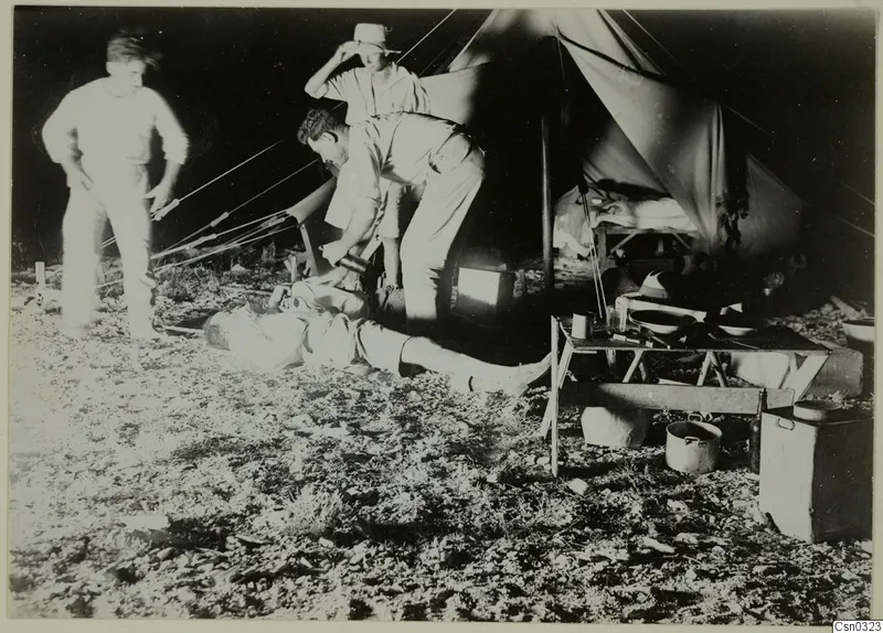 Three men by a canvas tent at night, with a folding table, enamel bowls and cooking pots on the ground