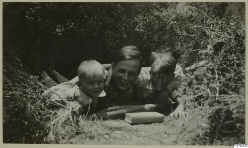 Einar Gjerstad and his two children lying in a gap in bushes, with a book on the ground in front of them