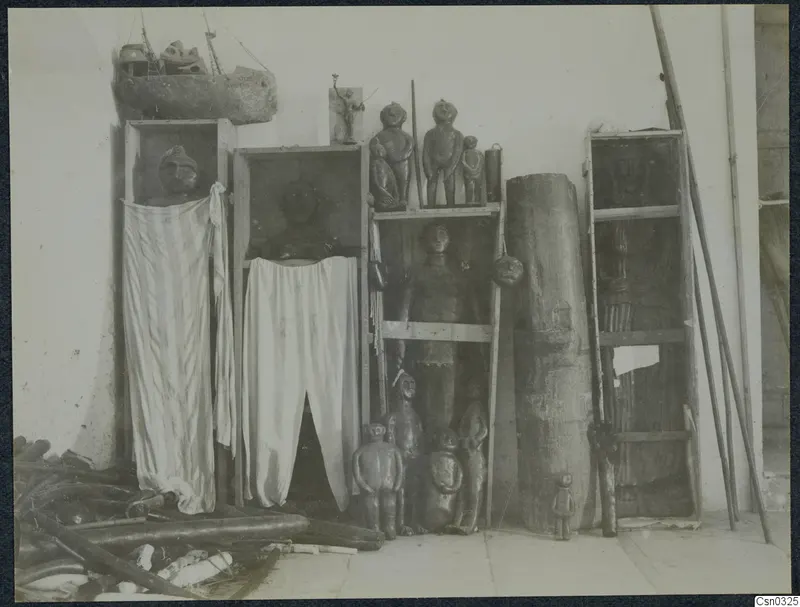 Carved wooden figures and draped cloths on wooden shelves and in open cabinets against a white wall