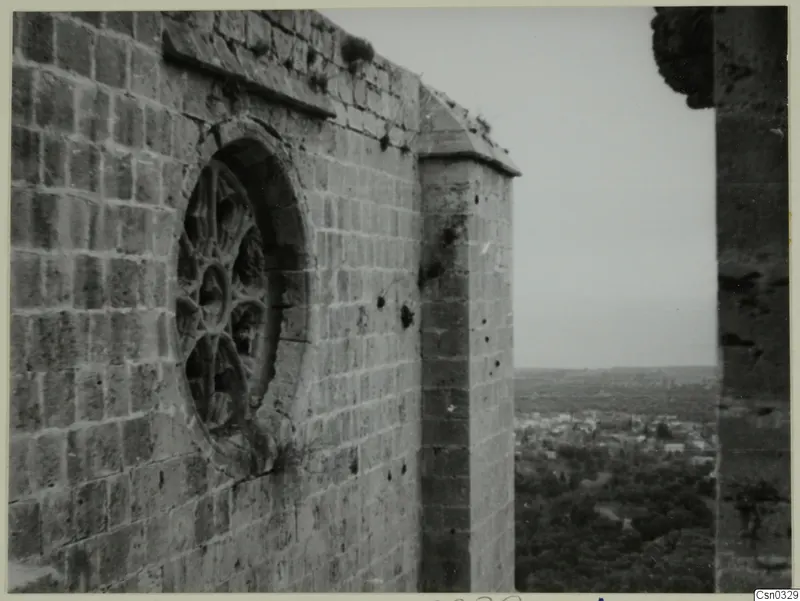 Stone wall with a round tracery window and buttress, with a view over a town and trees below, at Vessberg