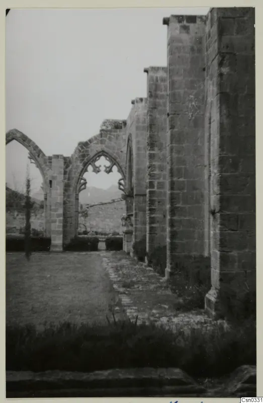 Ruined monastery walls with pointed arches and a paved path beside a grassy courtyard