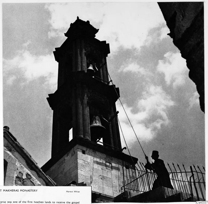 Belfry of Makheras Monastery seen from below, with a man holding a rope beside an iron railing.