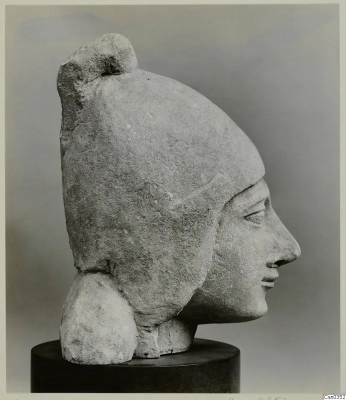 Limestone head of a figure in profile, wearing a tall headdress, shown on a dark cylindrical stand.