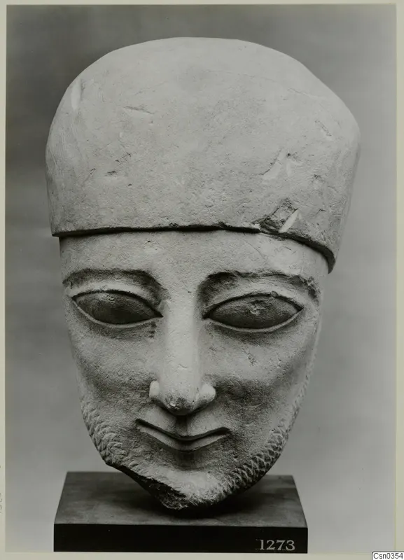 Limestone head of a bearded figure with a tall flat-topped cap, standing on a dark square base.