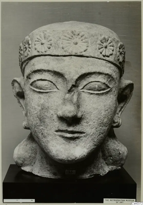 Frontal view of a carved limestone head with a rosette-decorated headband and large ears, on a black stand