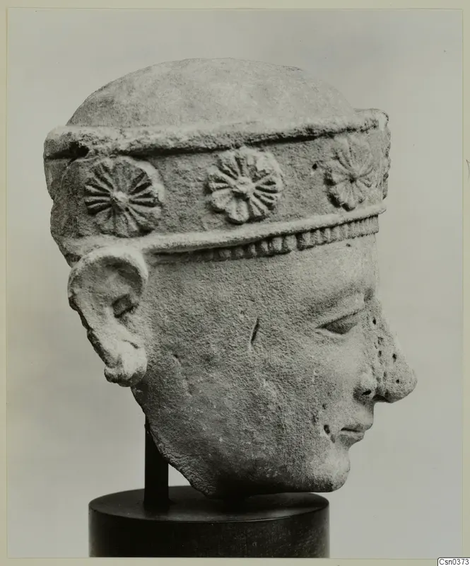 Carved limestone head of a figure wearing a band with rosette ornaments, shown on a dark stand