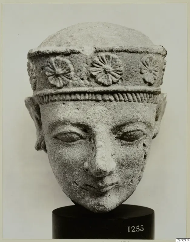 Limestone head of a figure wearing a band with rosettes, on a black stand with a number label.