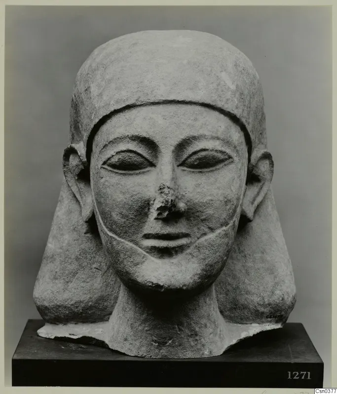 Carved limestone head with a close-fitting headdress and broad ears, nose broken, set on a dark wooden base