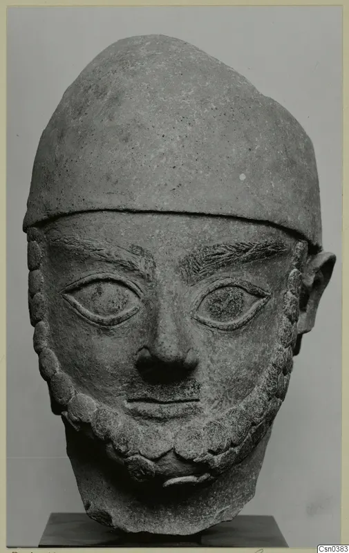 Carved stone head of a bearded man in a close-fitting cap, shown frontally on a dark stand.