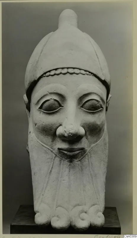 Limestone sculpture of a head with a tall rounded headdress, wide almond-shaped eyes and a broad nose, standing on a dark plinth
