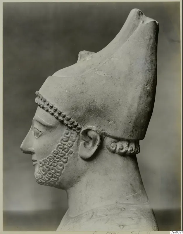 Limestone sculpture of a bearded head in profile, facing left, wearing a tall headdress with a curled neck band.
