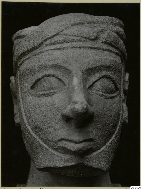Carved limestone head with a banded headdress and broad face, seen from the front against a dark background