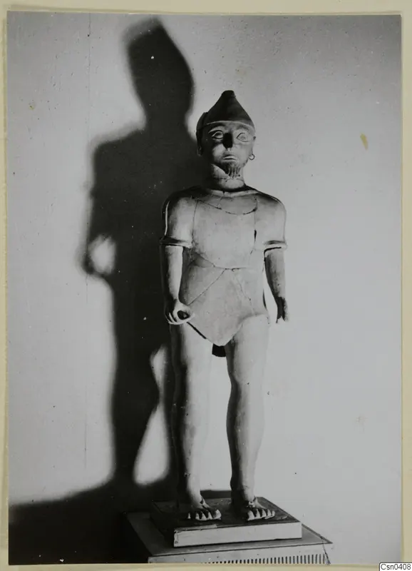 Standing terracotta statue of a figure in a pointed cap and short-sleeved tunic, set on a stepped base against a wall.