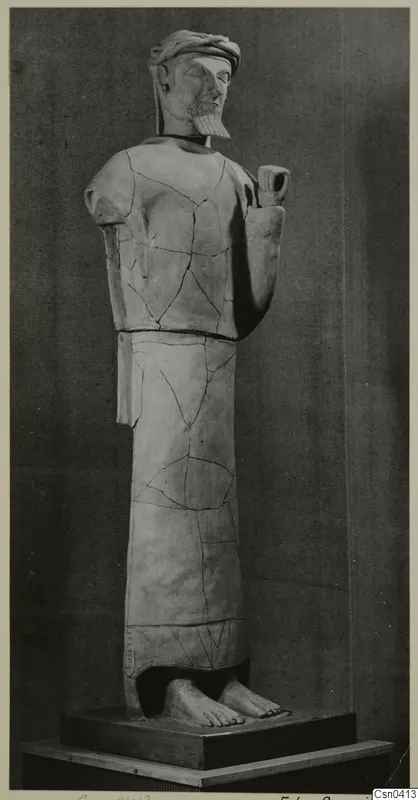 Standing male terracotta statue with a headband, beard and raised right hand, shown on a dark plinth against a grey backdrop.