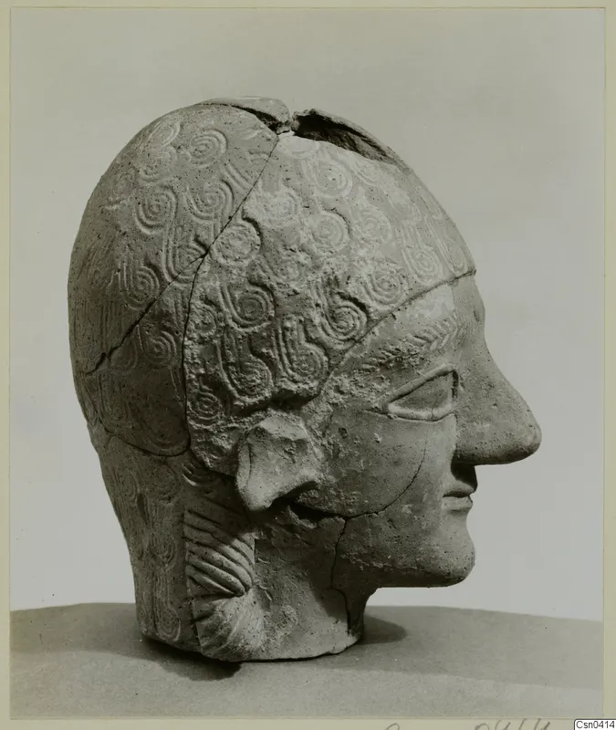 Terracotta head in side profile, with incised spiral pattern on the hair and a split, broken crown