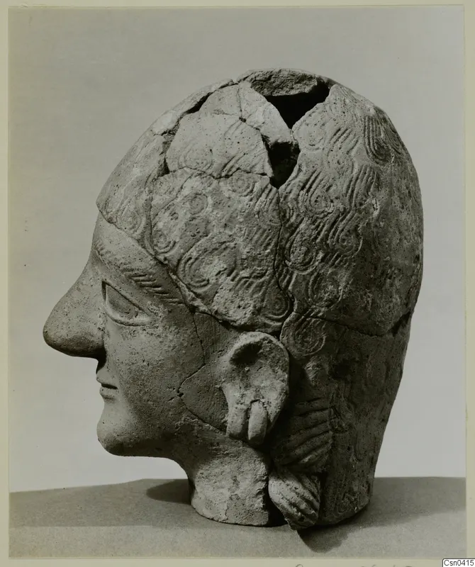 Terracotta head in profile facing left, with incised wavy hair and the top of the skull broken open.