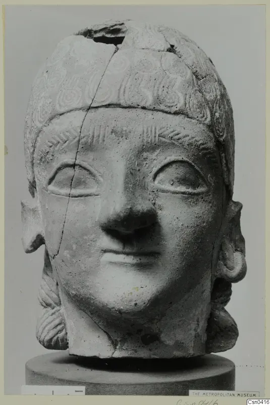 Terracotta head with incised curled hair and a broken crown, shown on a grey cylindrical stand