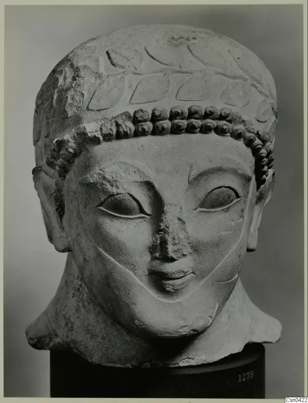 Limestone head of a figure with a laurel-like band of leaves and rows of curls over the forehead, photographed against a grey backdrop.