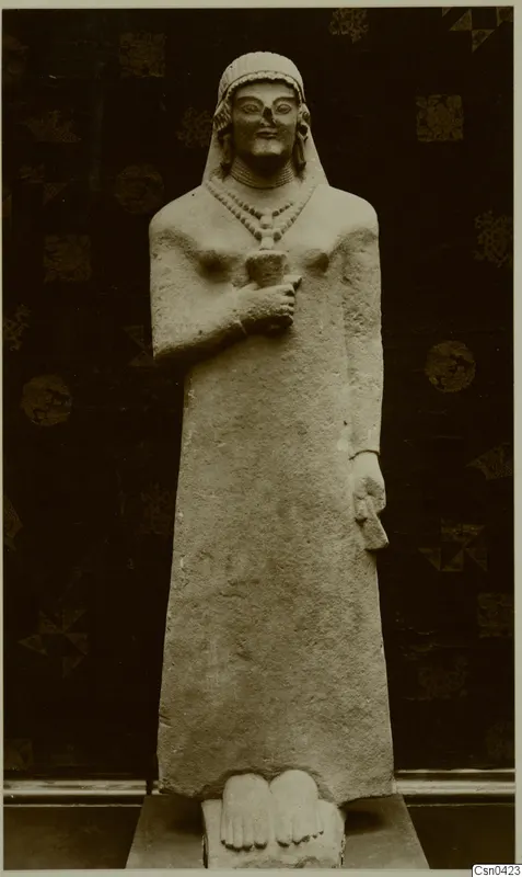 Limestone standing statue of a figure with a close-fitting head covering and beaded necklace, photographed against a dark patterned cloth.