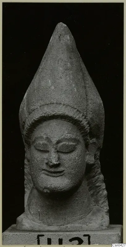 Limestone head statue with a tall conical headdress and closed eyes, shown against a black background.