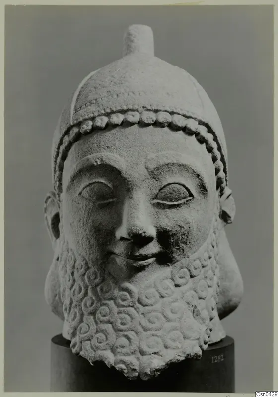 Limestone head of a bearded sculpture wearing a cap, on a dark cylindrical base