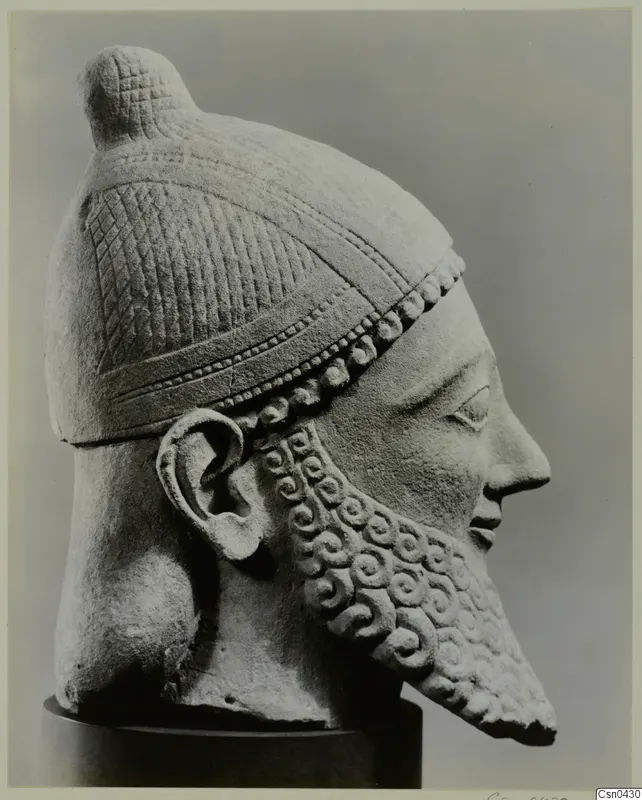 Limestone head of a bearded male figure in a helmet with a knob crest, shown in profile to the right.