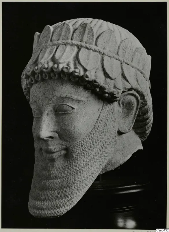 Limestone head of a statue with a leafy or petal-patterned headdress and a long, textured beard.