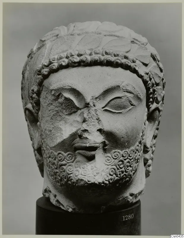 Limestone sculpture head of a bearded man, with curled hair and beard; the nose and part of the face are broken.