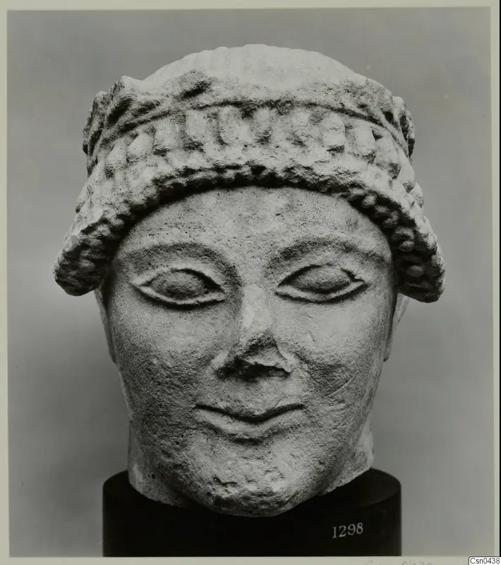 Limestone head of a figure with a banded headdress and almond-shaped eyes, shown on a black stand.