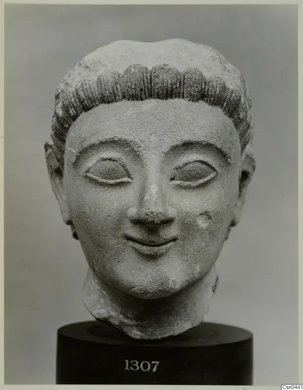 Limestone sculpted head of a figure with rows of curled hair and a faint smile, on a dark base.