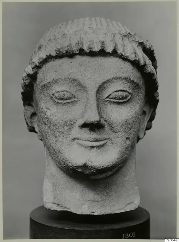 Limestone head of a figure with grooved hair, eyes and smiling mouth, shown on a dark cylindrical base.