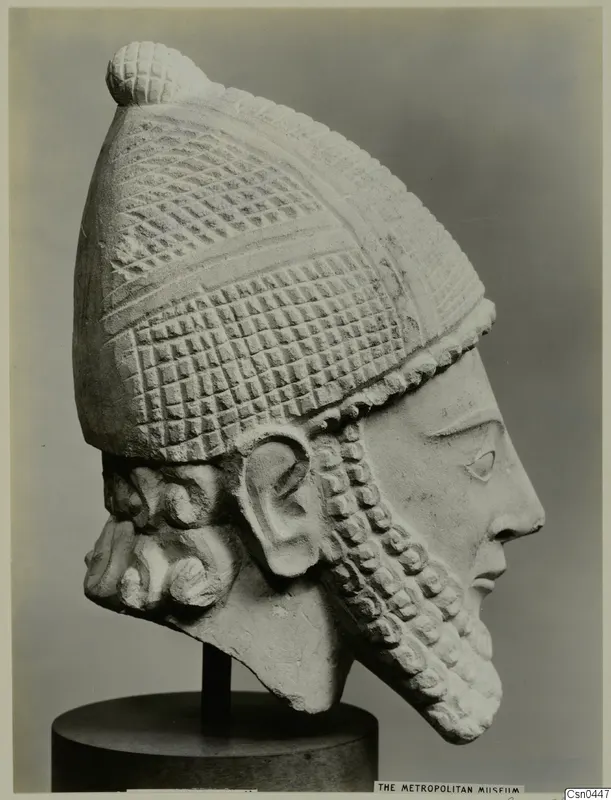 Limestone head of a bearded figure in a tall, grid-patterned helmet, shown in profile on a dark stand.