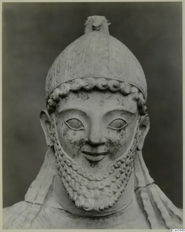Limestone sculpture of a bearded male head in a helmet with a crest, photographed against a dark backdrop.