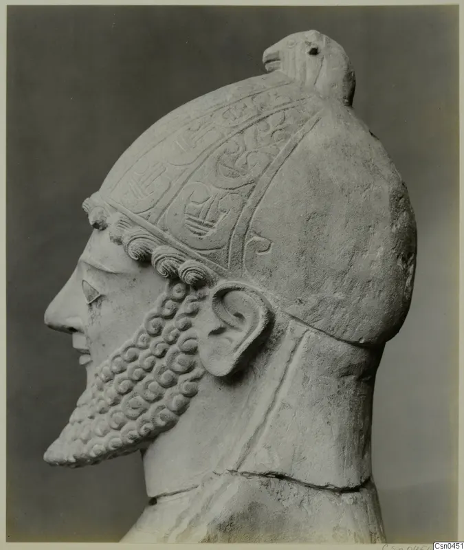 Limestone sculpture of a bearded head in profile, wearing a helmet with incised decoration and a bird-like crest.