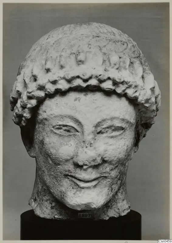 Limestone head of a figure with a curled hair band, photographed against a grey backdrop on a black stand.
