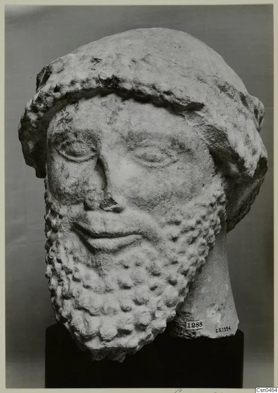 Limestone head of a bearded male sculpture with a wide band over the hair, shown against a grey backdrop.
