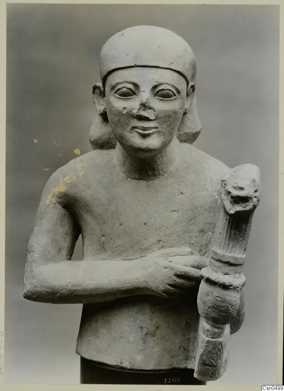 Bust-length limestone sculpture of a figure in a close-fitting cap, one arm across the chest, gripping a fluted upright object