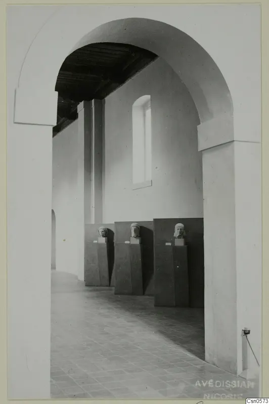 Three sculpted heads on dark pedestals in a museum gallery, seen through a tall arched doorway.