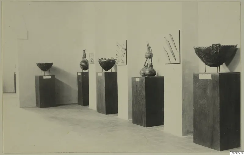 Five objects on pedestals in a gallery: bowls, a tall-necked flask, a stacked group of vessels, and blade-like tools on the wall