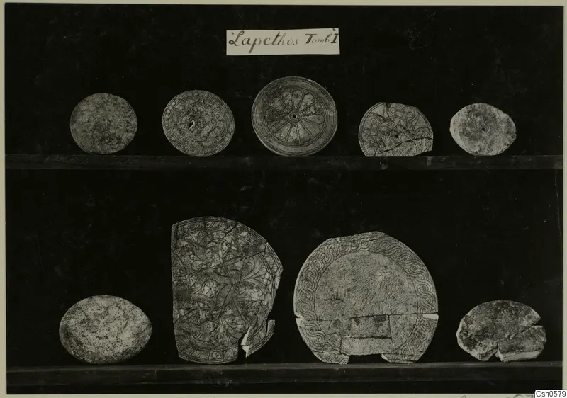 Nine round objects from Lapithos, Aj. Anastasia, T.I, some with patterns, shown on two shelves against a dark background.