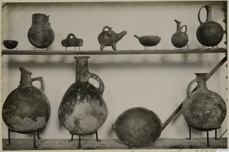 Two wooden shelves of pottery vessels on iron stands, mostly jugs and juglets, with a few bowls.