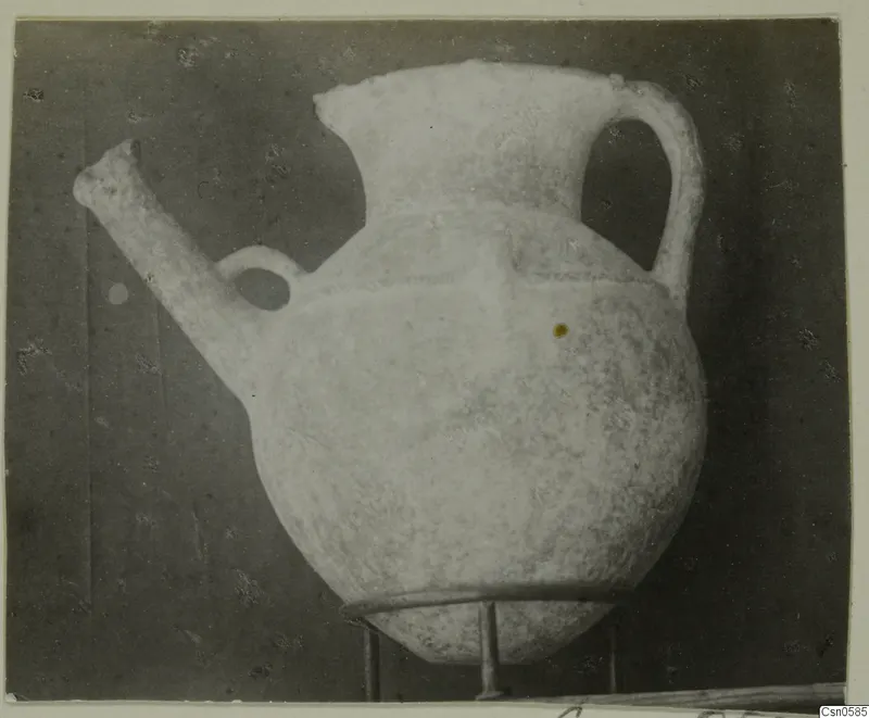 Pale pottery jug with a long spout, a small loop handle and a tall strap handle, shown on a metal stand