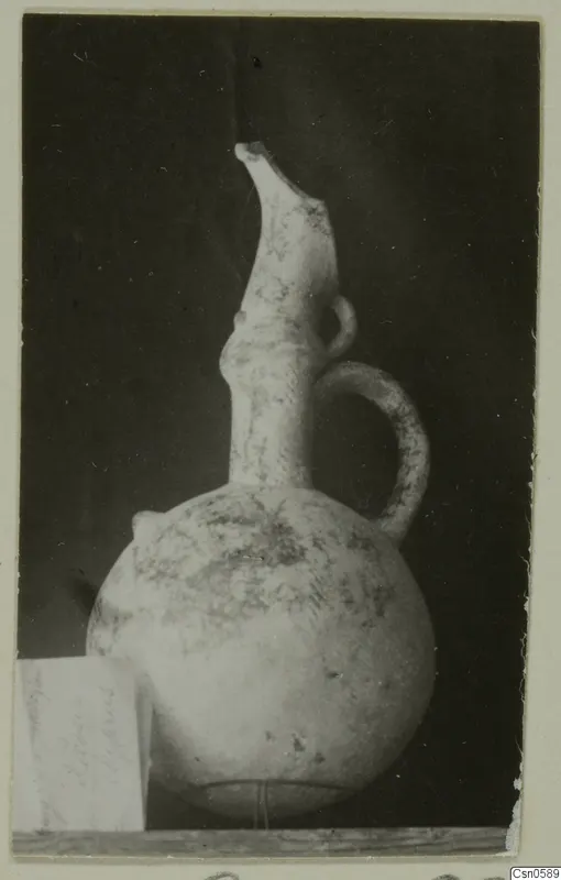 Pottery vessel with a globular body, tall flaring spout and strap handle, photographed on a wooden shelf